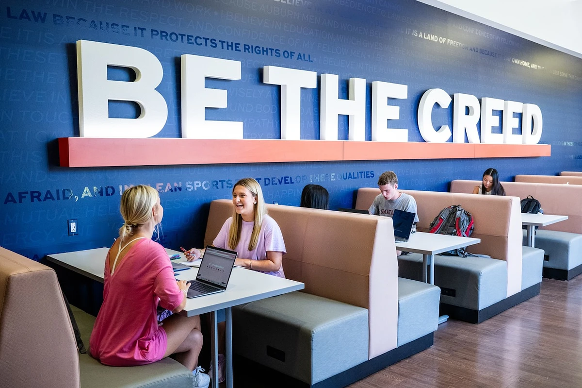 People are sitting in a modern study lounge with a large "BE THE CREED" sign on the wall.