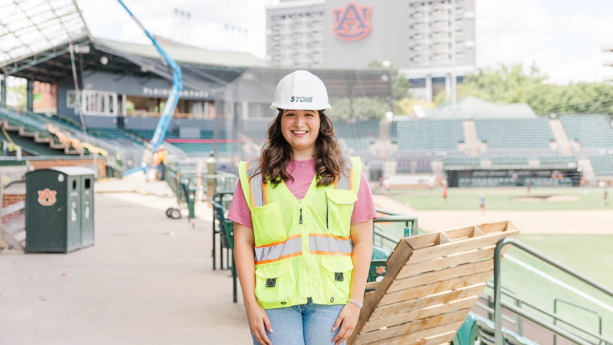 A woman wearing a hard hat and high-visibility vest stands smiling in a stadium under renovation.