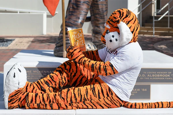 A person in a tiger costume is sitting and reading a book titled "The Music of Bees."