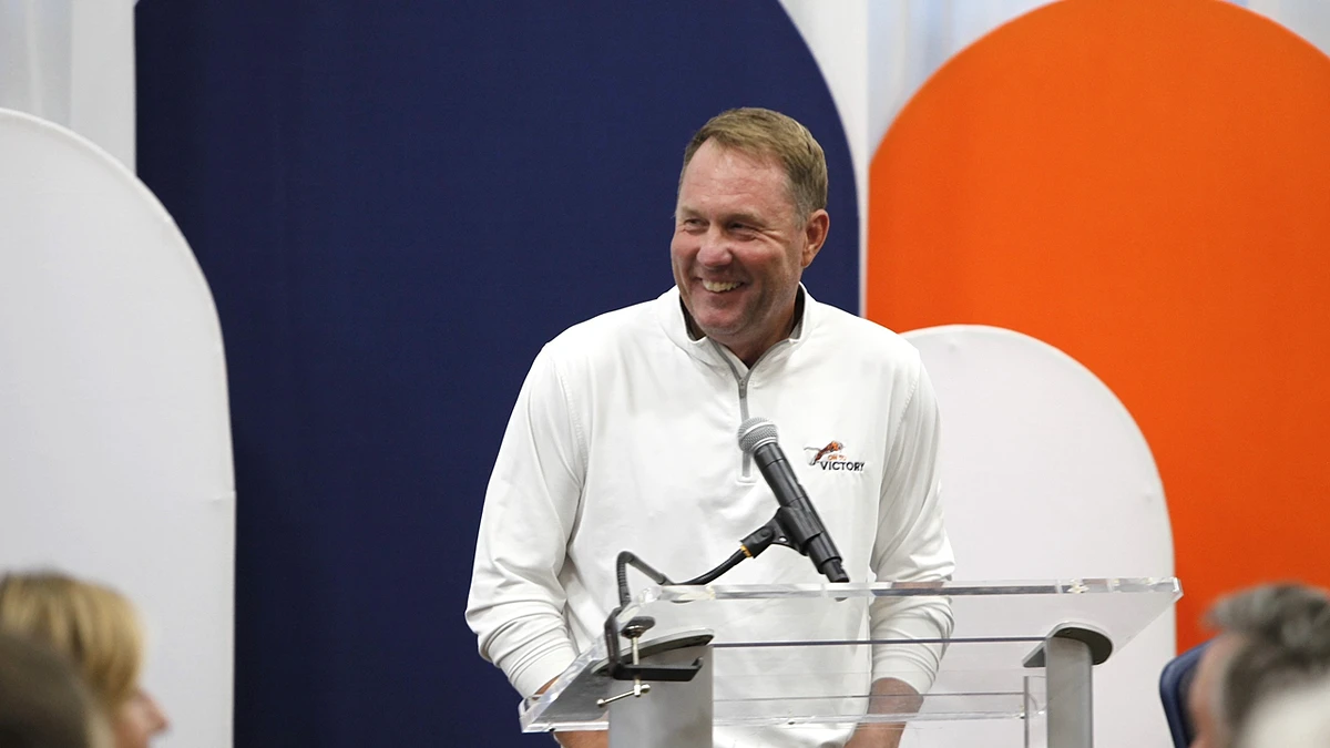 A man wearing a white pullover stands behind a podium and microphone, smiling during a speech, with colorful background panels behind him.