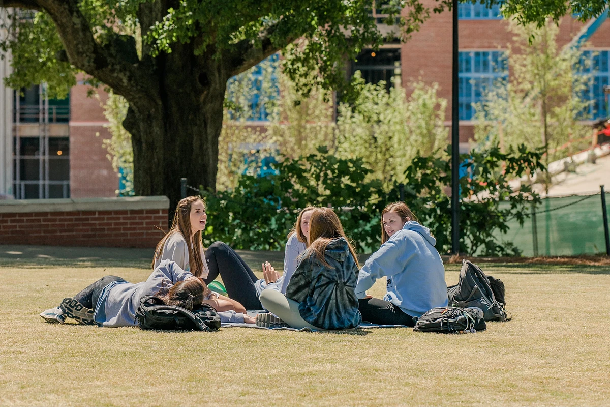 A group of people are sitting and chatting on a grassy area under a tree with a building in the background.