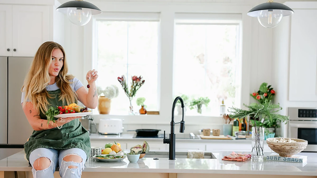 A woman holding a plate of food sits on a kitchen counter decorated with fresh produce and flowers.