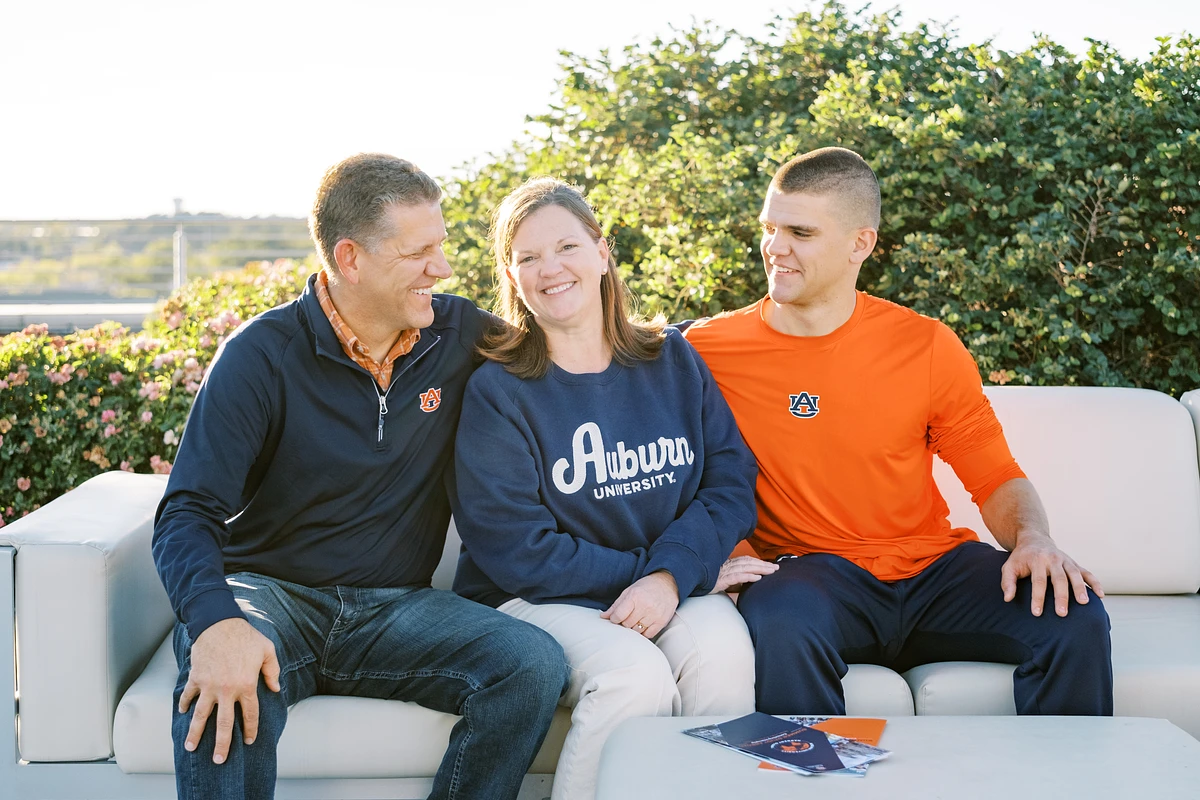 Three people wearing Auburn University clothing are sitting together on an outdoor couch, smiling.