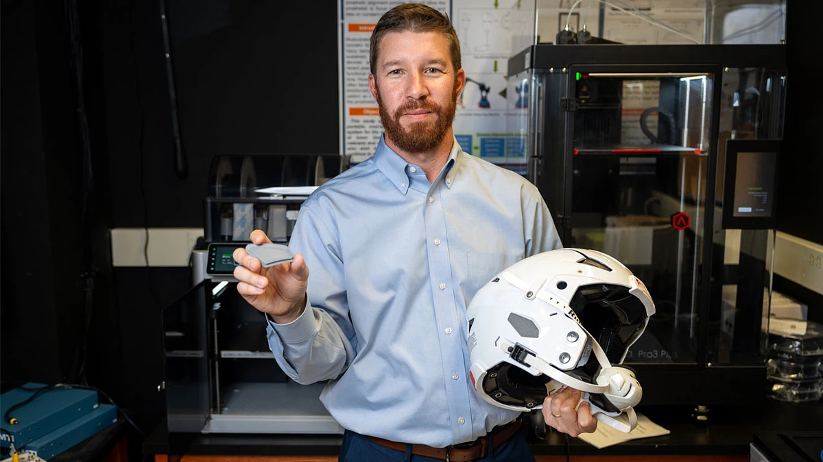 A man in a lab setting holds a white helmet and a small object.