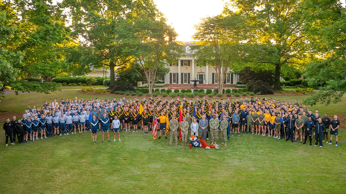 A large group of people, dressed in various uniforms, pose together on a lawn in front of a large house surrounded by trees.