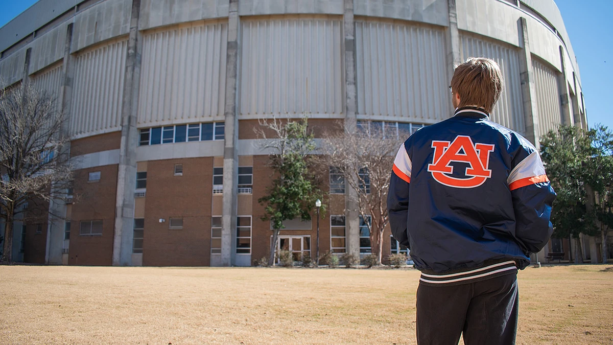 A person wearing a jacket with "AU" stands in front of a large building.