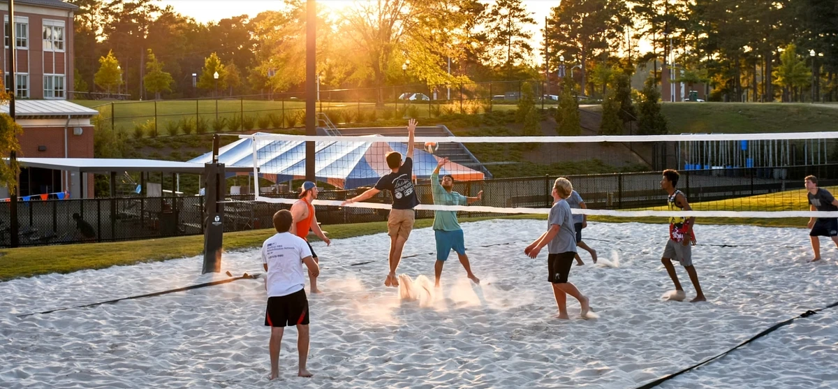 A group of people is playing beach volleyball on a sand court during a sunny evening.