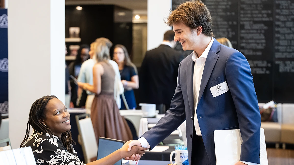 A man in a blue suit is smiling and shaking hands with a seated woman at a professional event.