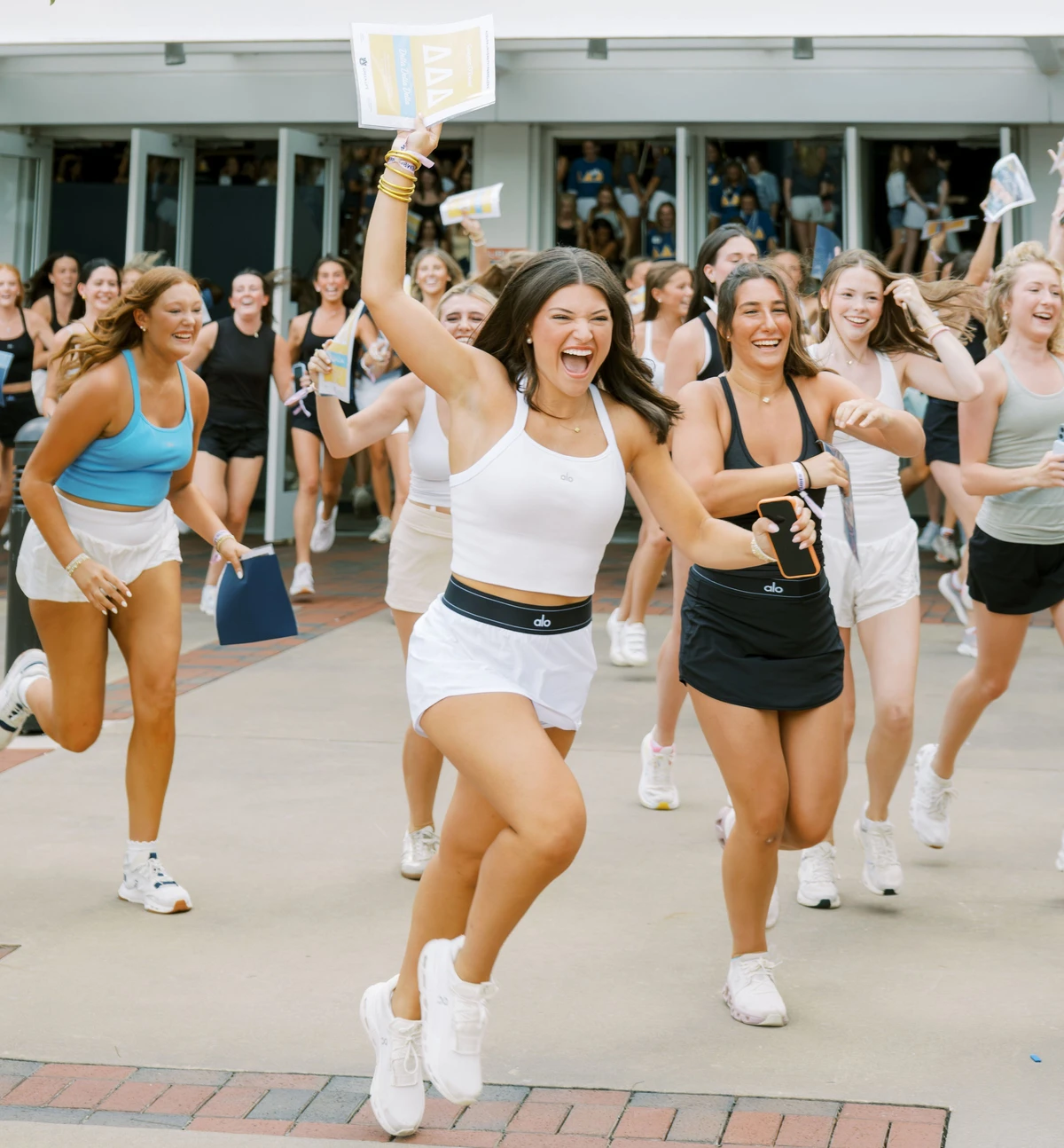 A group of women run down a street, smiling and holding papers.